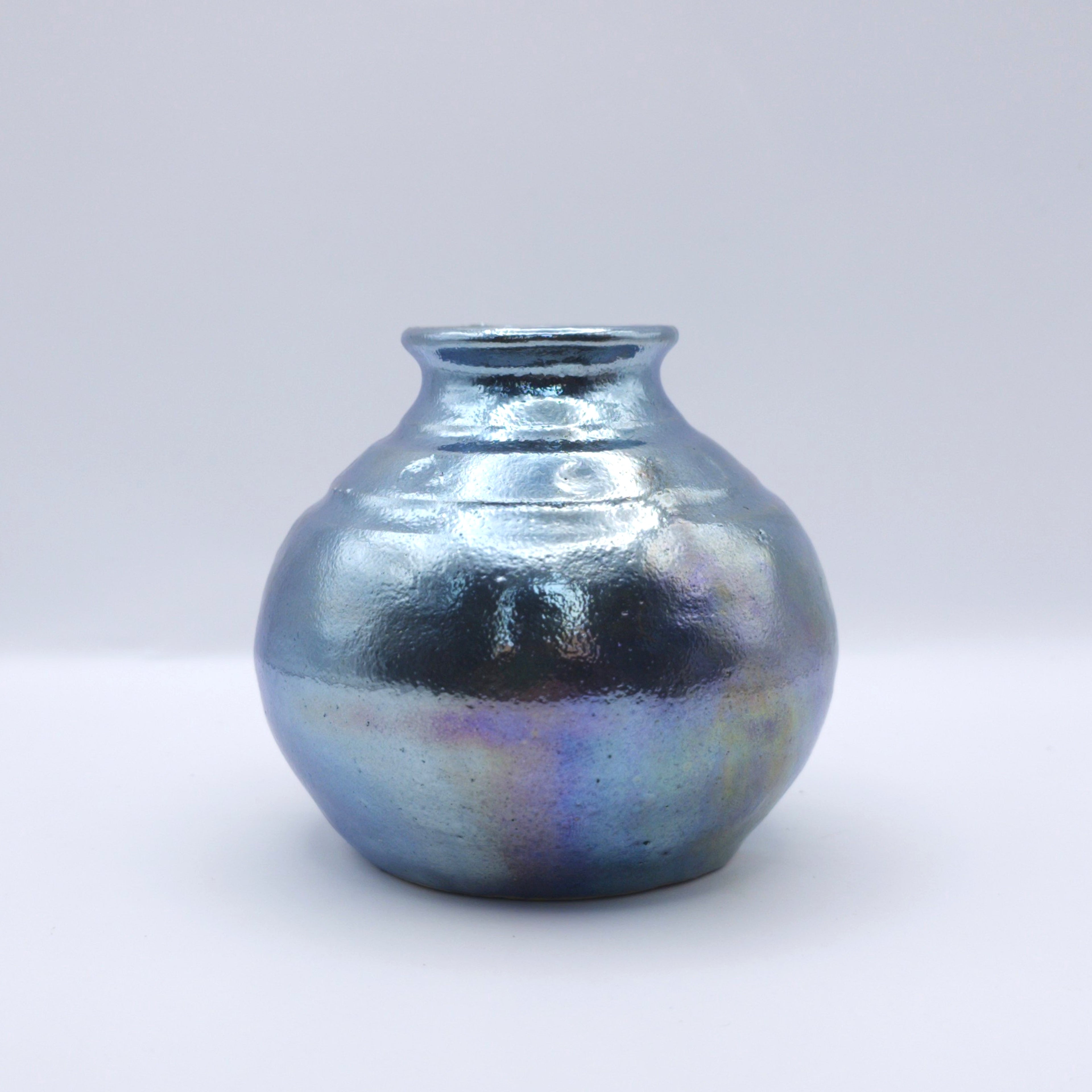 Pequeña vasija cerámica de forma redondeada con acabado metálico e iridiscente. La superficie refleja tonos azules, violetas y plateados que cambian según la luz, creando una apariencia luminosa y experimental.