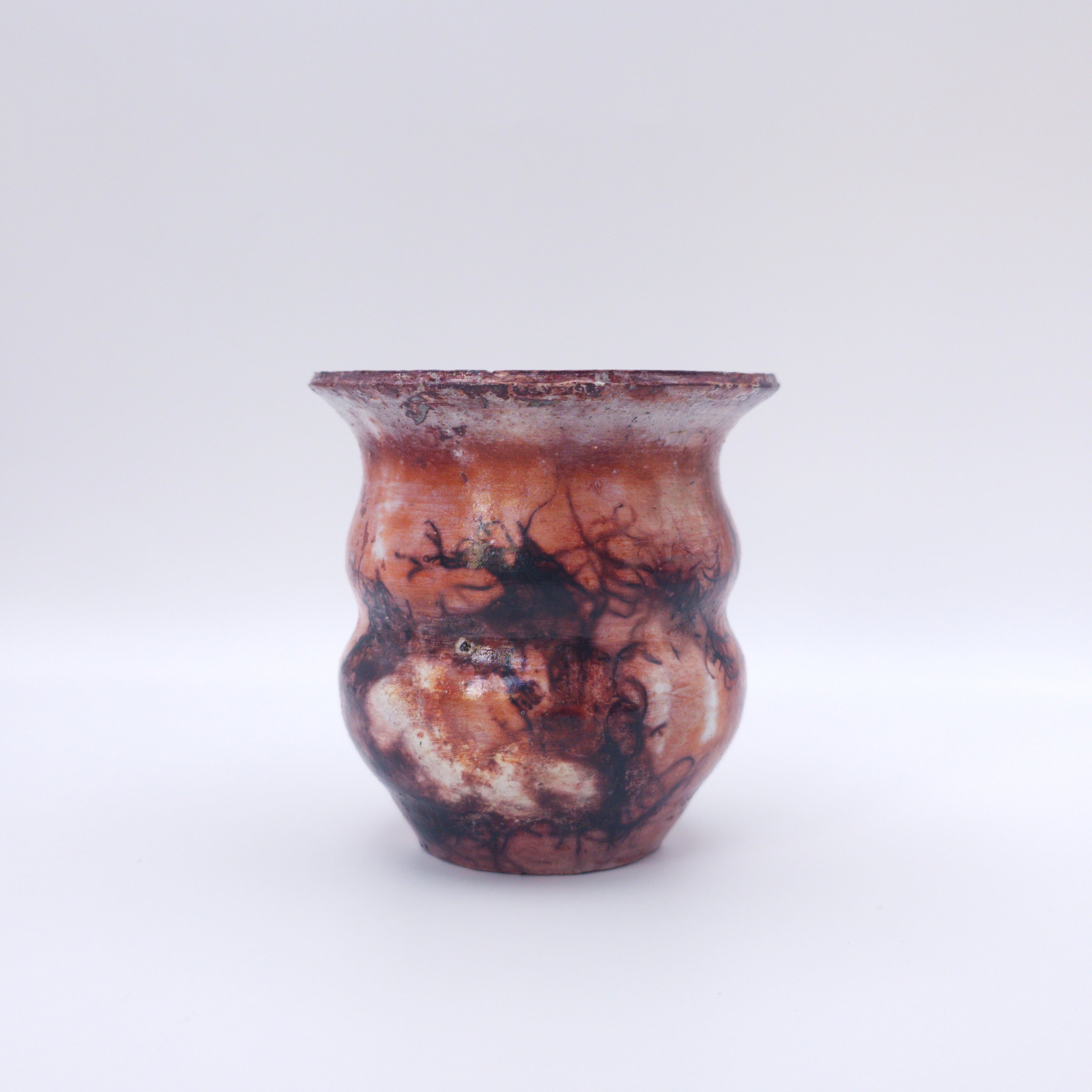 Pequeña vasija cerámica de forma orgánica con superficie en tonos marrones, rojizos y tierra. La pieza muestra marcas oscuras ramificadas producidas por la cocción saggar con cloruro férrico, creando una textura experimental inspirada en fuego, humo y materia natural.